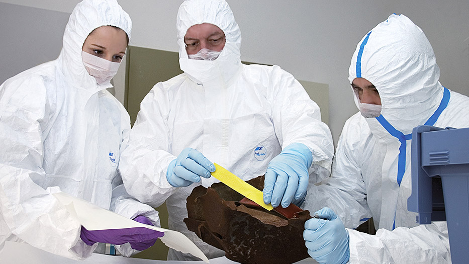 drei Personen in Schutzkleidung und Mundschutz bei der Spurensicherung an einem Asservat (verweist auf: Spurensicherung im Labor)